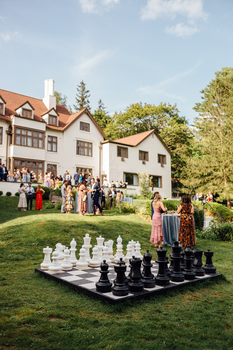 A large outdoor chessboard with oversized black and white pieces sits on a grassy lawn at a Seven Hills Inn Wedding in Lenox, MA. Two women stand by a cocktail table as a crowd gathers outside the grand white house in the background.