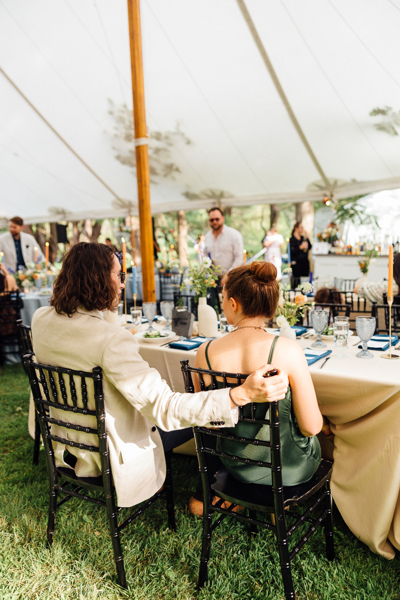 A man with long hair and a light jacket sits with his arm around a woman in a green dress at a Seven Hills Inn wedding in Lenox, MA, under a white tent surrounded by tables, chairs, and guests.