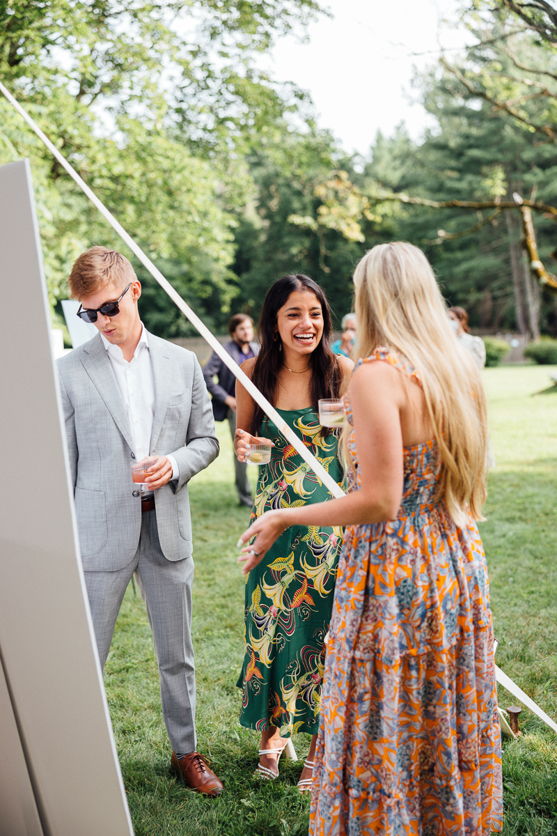 Three people stand outdoors on grass, chatting and smiling at a Seven Hills Inn Wedding in Lenox MA. One man in a grey suit and sunglasses holds a drink, while two women in colorful dresses laugh. Trees and other guests are visible in the background.