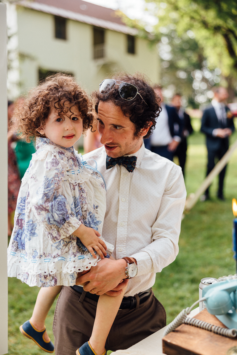 At a Seven Hills Inn Wedding in Lenox MA, a man in a bow tie and sunglasses on his head holds a young child in a floral dress outdoors amid guests and lush greenery.