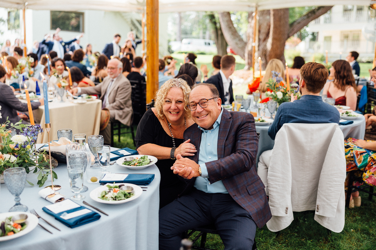 A smiling couple sits close together at a round table with salads and drinks at an outdoor Lenox MA event. Flowers and candles set a festive mood, suggesting a celebration or even a Seven Hills Inn wedding reception.
