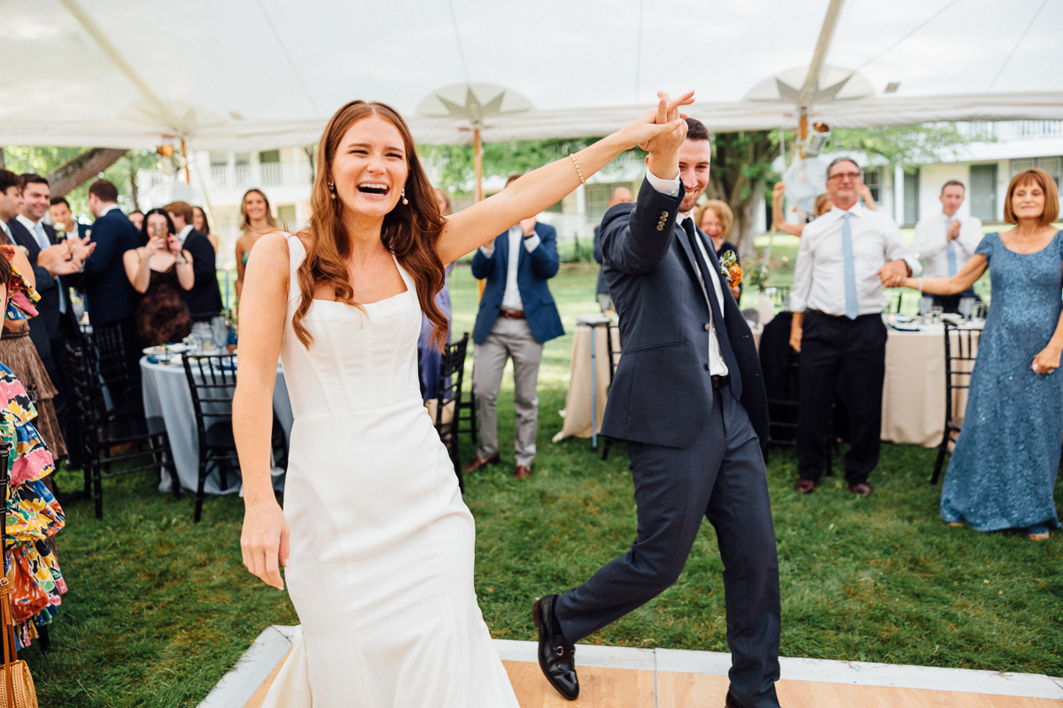 A smiling bride and groom hold hands and dance into their Seven Hills Inn Wedding reception in Lenox, MA under a white tent, surrounded by cheering and clapping guests.