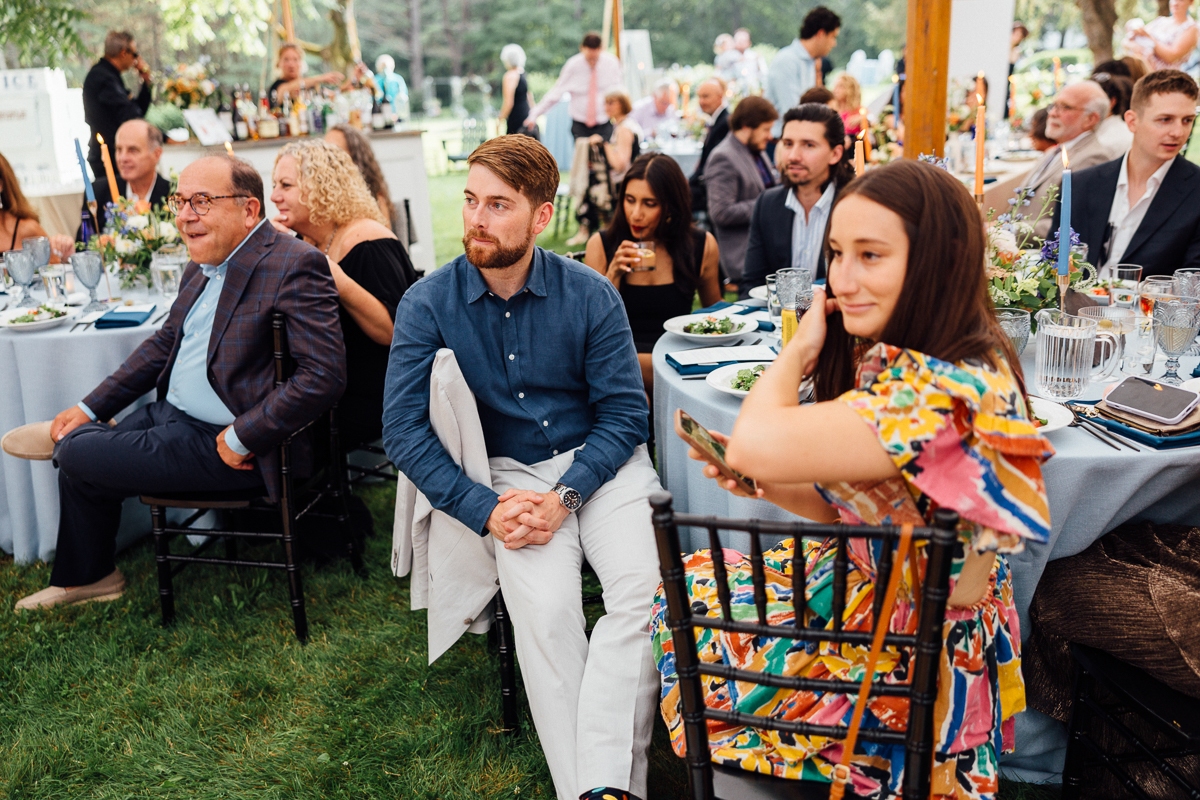 Guests seated at round tables outdoors during a Seven Hills Inn wedding in Lenox MA, listening attentively. The tables are covered with light blue tablecloths and decorated with flowers. People are dressed in semi-formal attire.