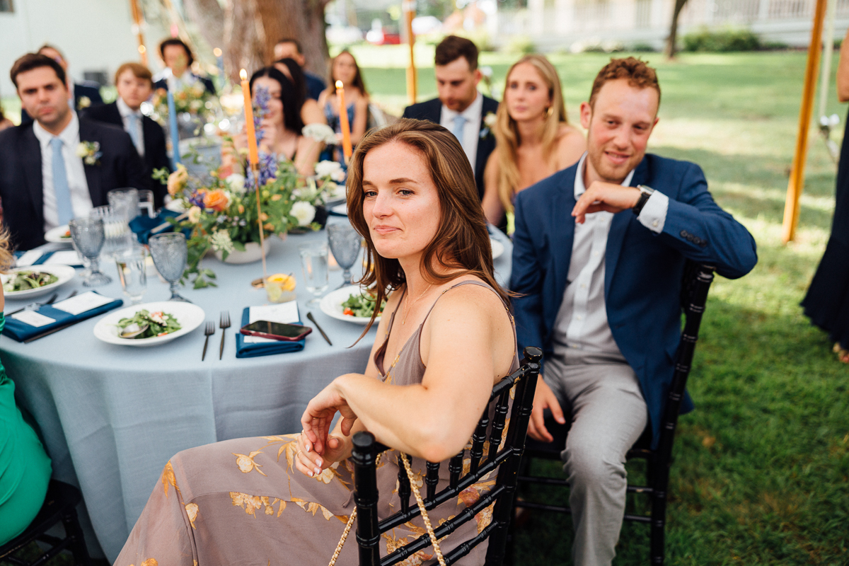 A woman in a floral dress sits at a round outdoor table, smiling slightly at the camera. Other guests in formal attire gather around her with food and flowers on the table at a Seven Hills Inn wedding in Lenox, MA.