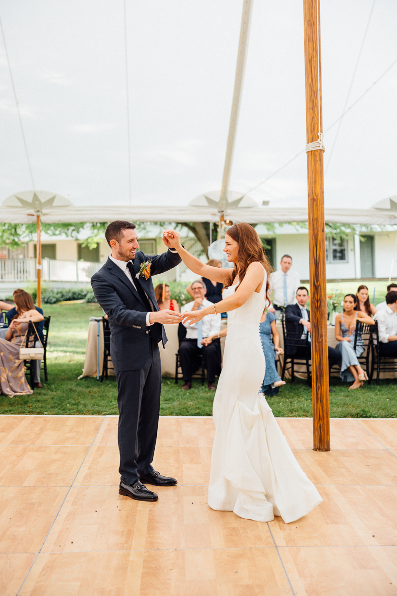 A bride and groom dance together on a wooden floor under a tent at their Seven Hills Inn wedding in Lenox, MA, surrounded by seated guests watching and smiling.