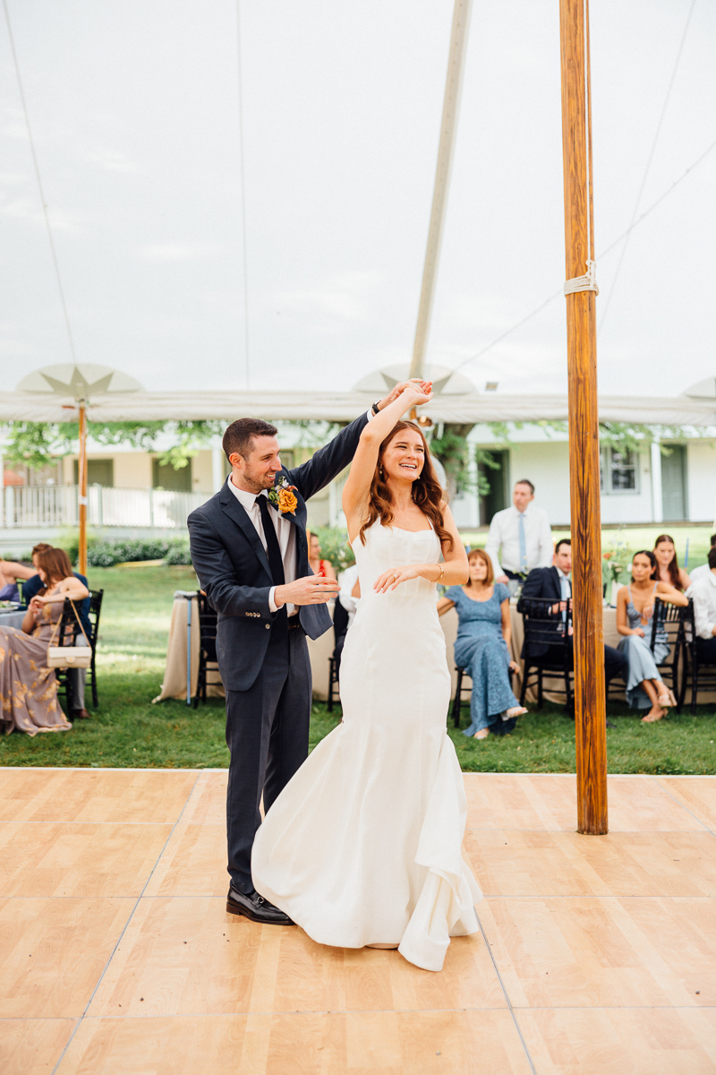 A bride in a white dress and a groom in a dark suit share a joyful first dance under a tent at their Seven Hills Inn wedding in Lenox, MA, surrounded by seated guests watching and smiling.