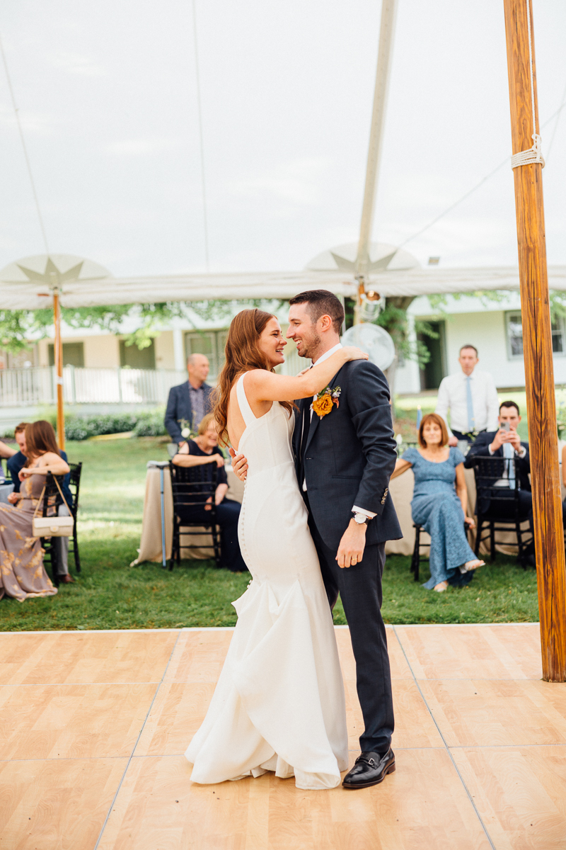 A bride and groom share a joyful first dance outdoors under a white tent at their Seven Hills Inn wedding, smiling at each other. Guests seated at tables in the background watch the couple on a wooden dance floor.