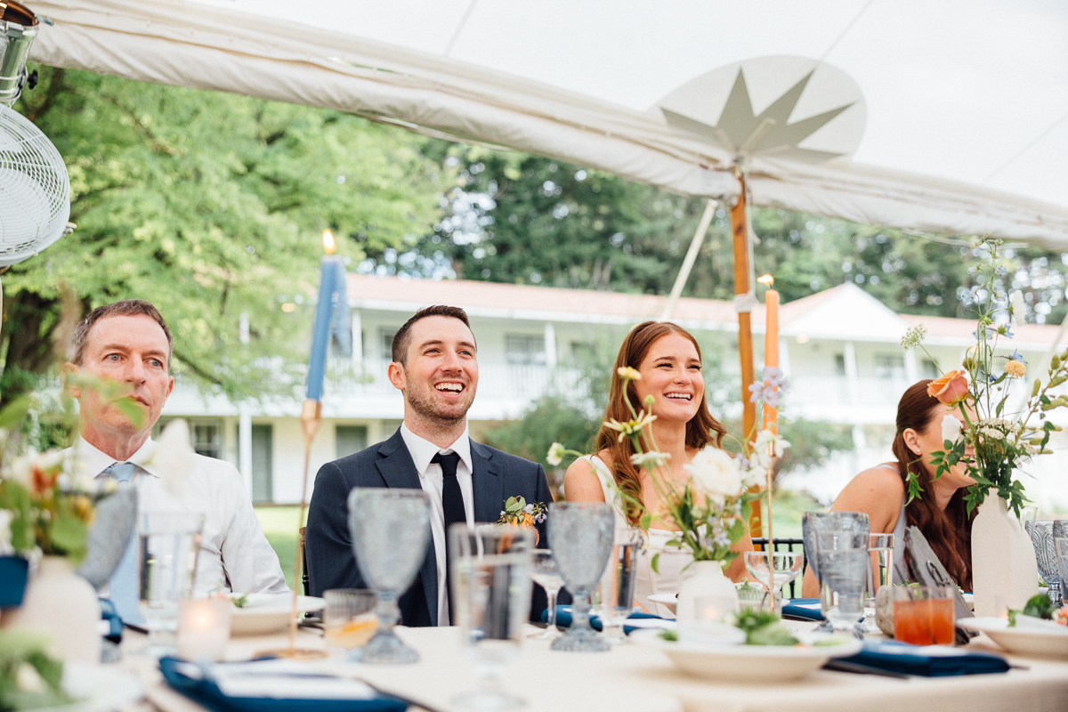 Four people dressed formally sit at a decorated outdoor table, smiling and enjoying a celebration. Set in lush greenery beneath a white tent, the scene captures the elegance of a Seven Hills Inn Wedding in Lenox, MA.
