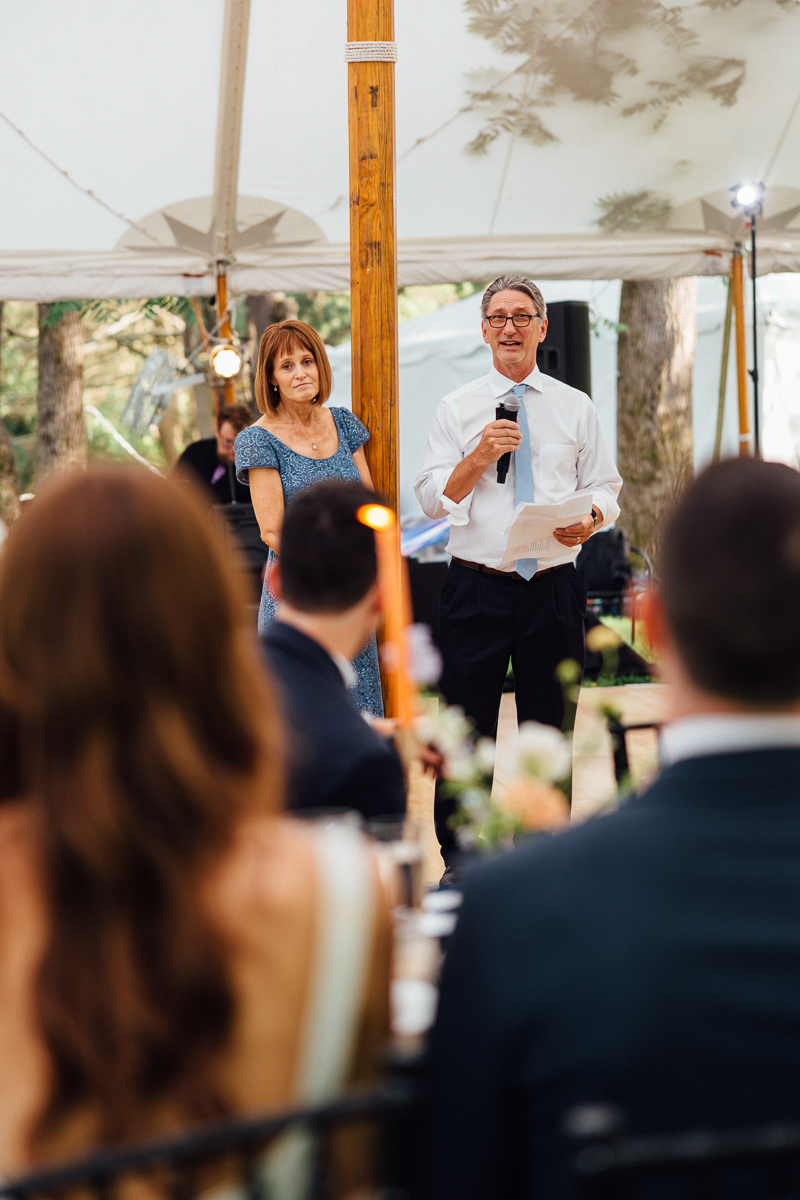 A man holding a microphone and paper stands next to a smiling woman under a tent, speaking to seated guests at a beautiful Seven Hills Inn Wedding in Lenox MA, with trees and string lights glowing in the background.