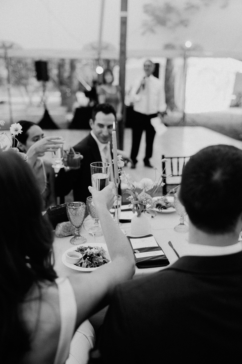 A black-and-white photo shows people raising champagne glasses in a toast around a table set with food and flowers, while a person speaks in the background. The celebratory atmosphere hints at a joyful Seven Hills Inn wedding in Lenox MA.
