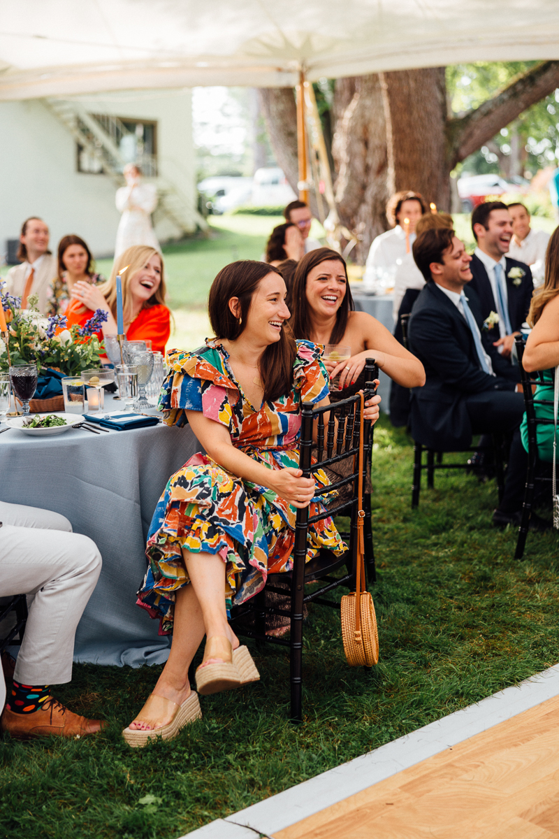 Two women sit smiling and laughing at an outdoor Seven Hills Inn wedding in Lenox, MA, surrounded by other guests at decorated tables. One wears a colorful floral dress and espadrilles, adding to the lively, festive atmosphere under the tent.