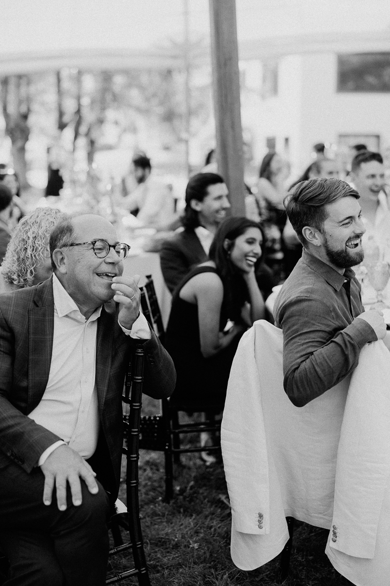 A group of people sit outdoors, smiling and laughing around tables at what appears to be a social or celebratory event in Lenox MA. The image, captured in black and white, evokes the timeless joy of a Seven Hills Inn Wedding.