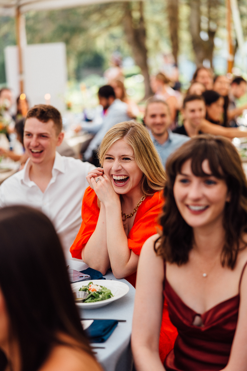 A group of people sit at a table outdoors, smiling and laughing at a Lenox MA wedding. The focus is on a woman in a bright orange dress, laughing with her hand near her face. Plates of food are on the table as others nearby join in the joy.
