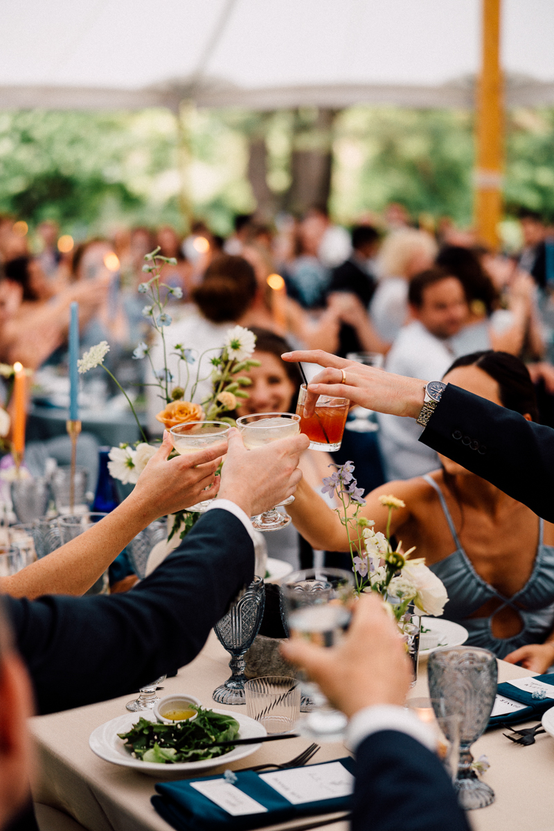 A group of people raise their glasses in a celebratory toast at an elegant Seven Hills Inn wedding in Lenox MA, seated around a decorated table with flowers, candles, and plates of salad. The scene is lively and festive.