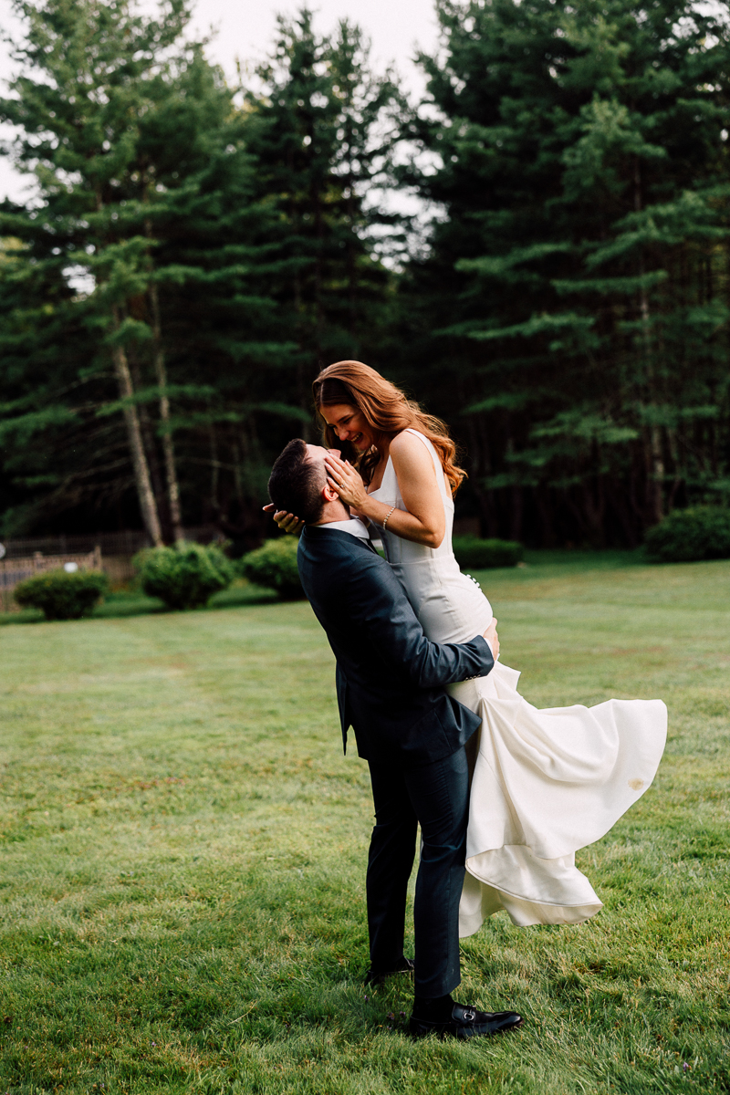 A groom in a dark suit lifts his bride in a white dress as they smile at each other on a grassy lawn, surrounded by tall trees during their romantic Seven Hills Inn wedding in Lenox, MA.