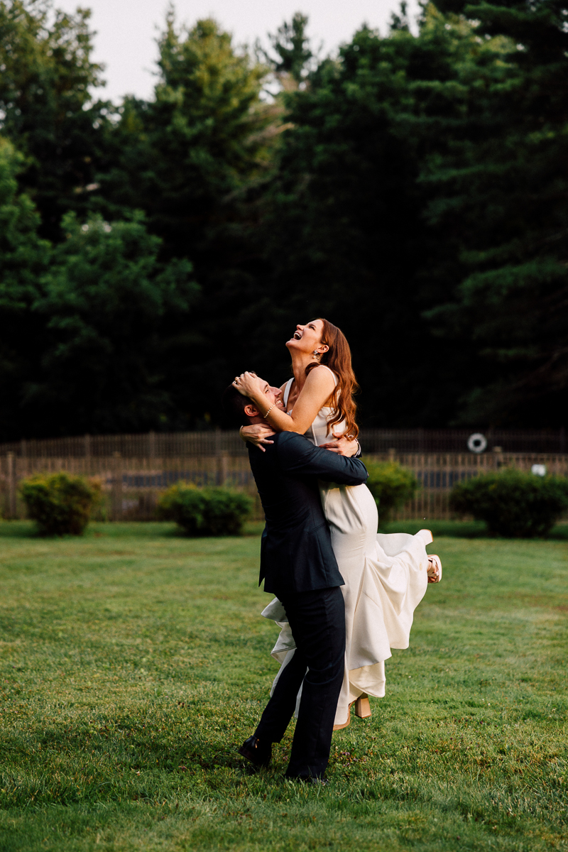 A man in a suit lifts a woman in a white dress as they smile and laugh together on the grassy lawn at their Seven Hills Inn wedding, with trees and a wooden fence providing a charming Lenox MA backdrop.