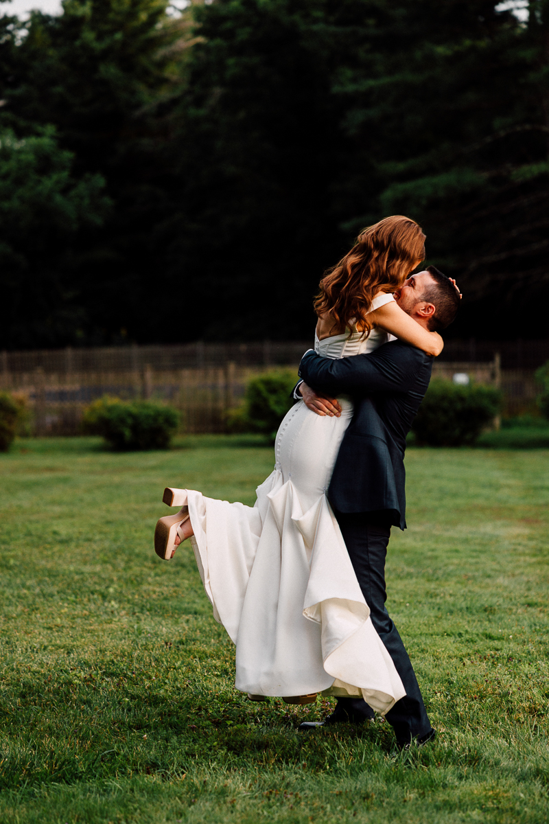 A man in a dark suit lifts a woman in a white wedding dress as they embrace outdoors on the grassy lawn of Seven Hills Inn in Lenox, MA, surrounded by trees, with the woman’s legs bent and her shoes visible.
