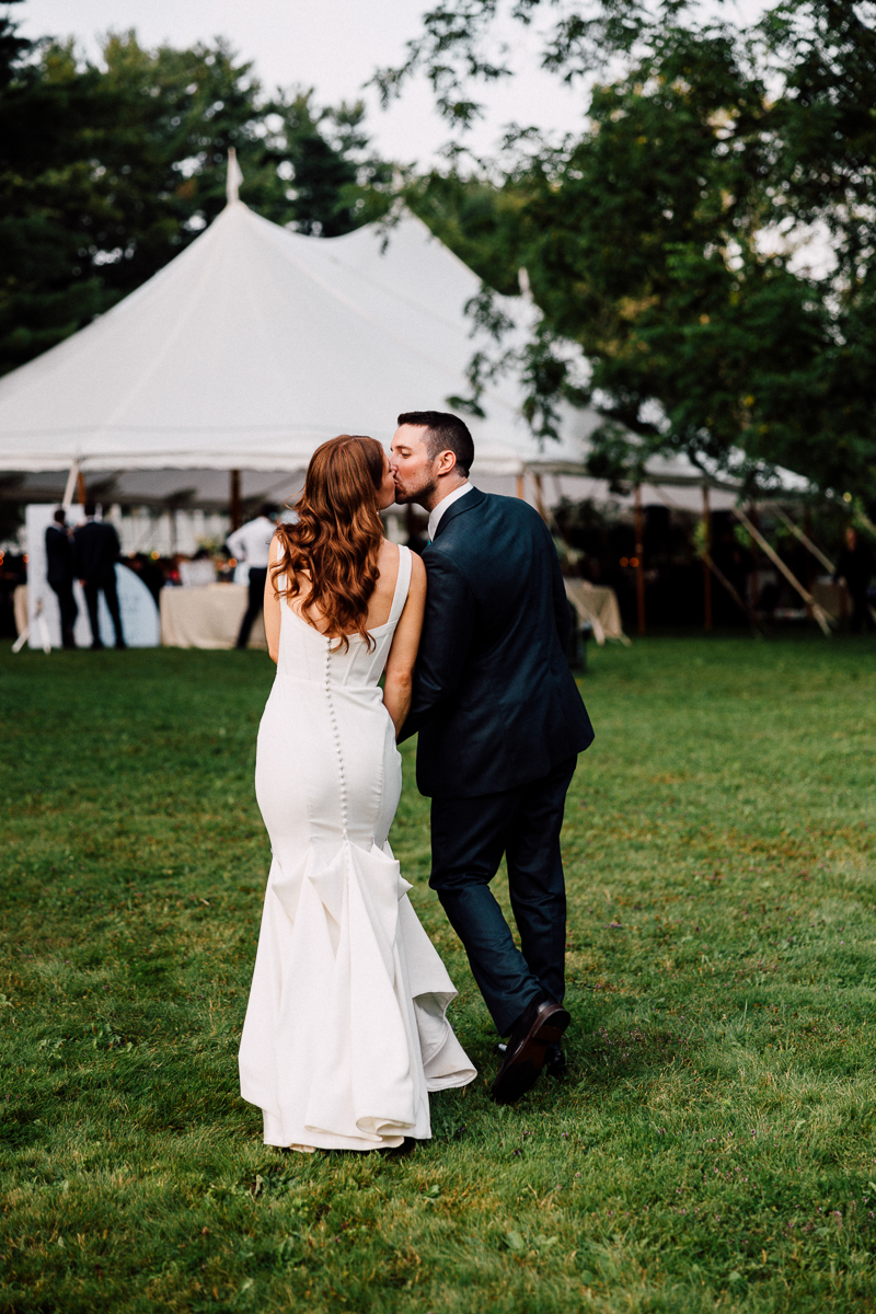 A bride in a white dress and groom in a dark suit walk hand in hand across a grassy lawn toward a white tent, surrounded by trees and guests at an outdoor Seven Hills Inn wedding in Lenox, MA.