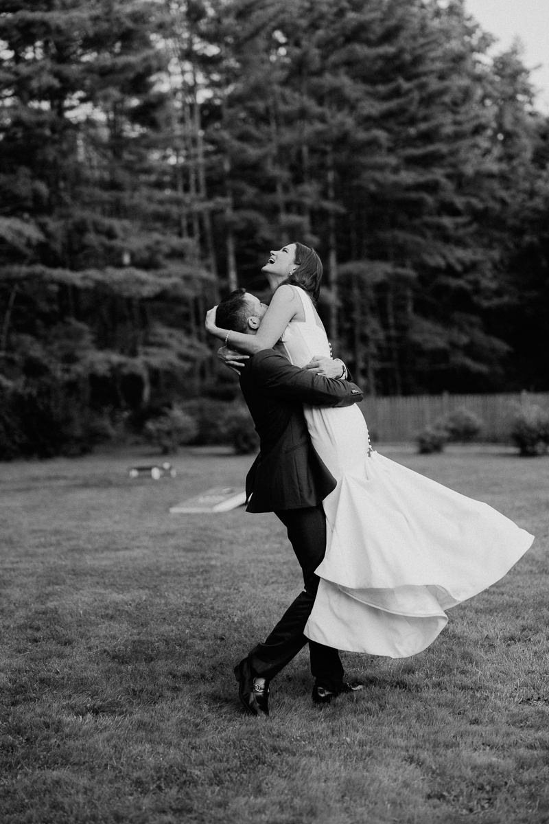 A groom lifts and spins his smiling bride in a joyful embrace outdoors at their Seven Hills Inn Wedding. The bride’s dress flows as they celebrate amid Lenox MA's tall trees and grass. The black-and-white photo captures pure happiness.