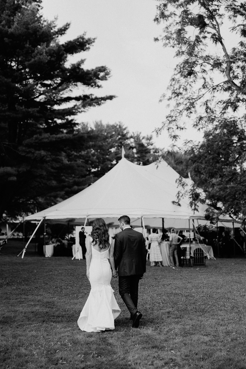 A bride and groom walk hand in hand toward a large outdoor tent at their Seven Hills Inn wedding reception in Lenox MA, surrounded by trees and guests, in a timeless black and white photo.