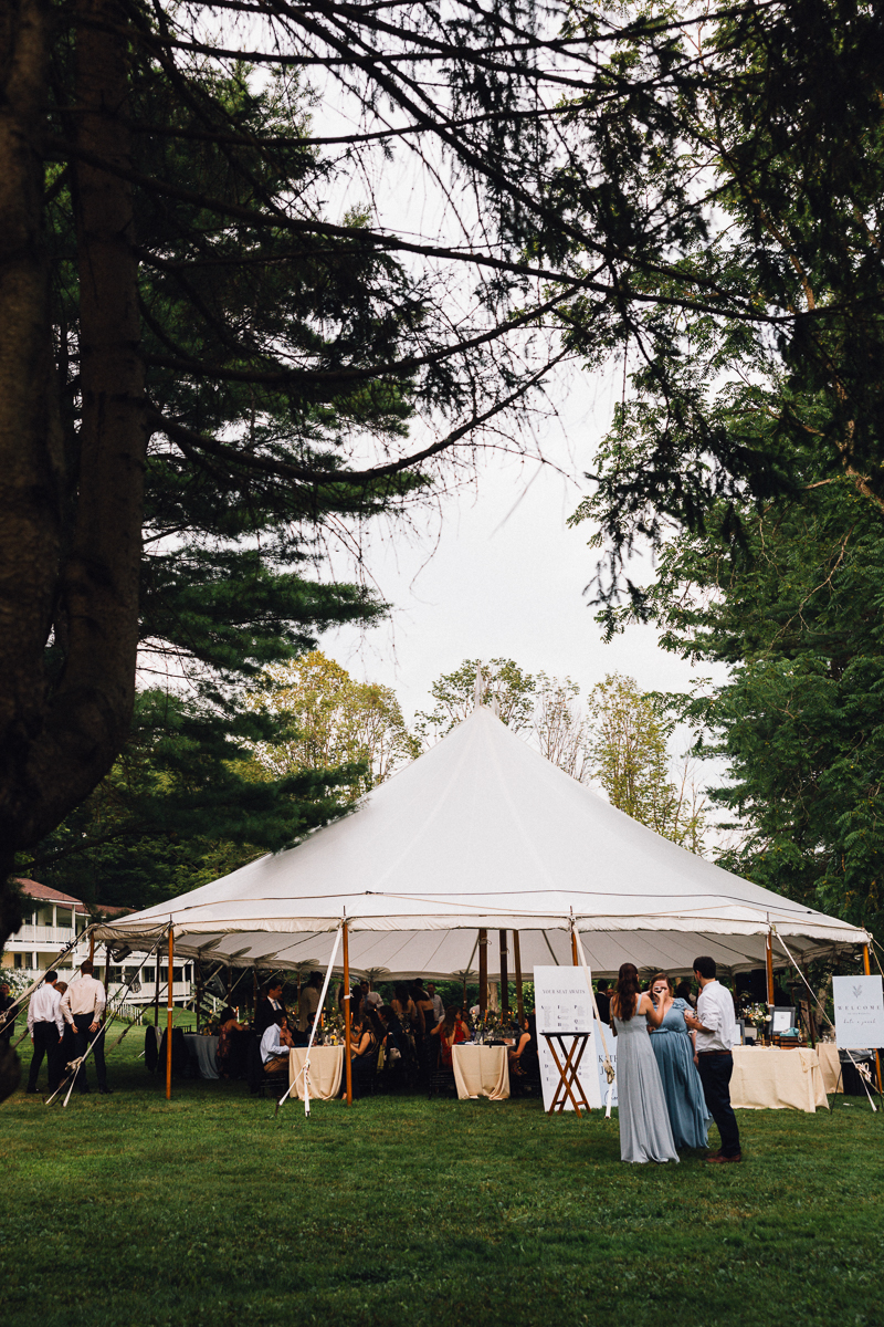 A large white tent set up outdoors on a grassy lawn at Seven Hills Inn in Lenox, MA, surrounded by tall trees. People in formal attire gather under and around the tent, suggesting a wedding or special event. Two women and a man stand in the foreground.