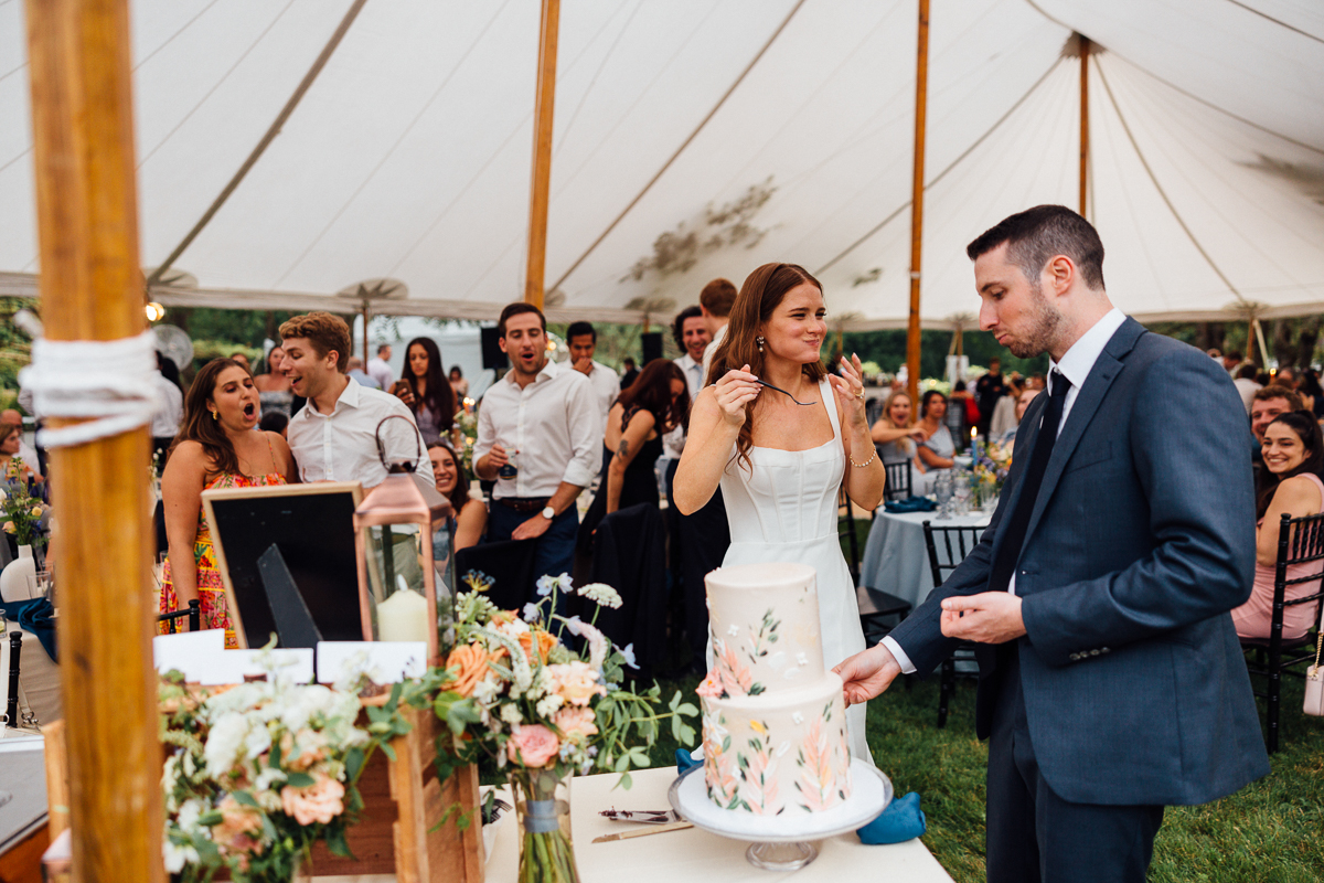 A woman in a white dress and a man in a suit stand by a decorated wedding cake under a large tent at their Seven Hills Inn wedding in Lenox MA, surrounded by guests celebrating amid flowers and party decorations.