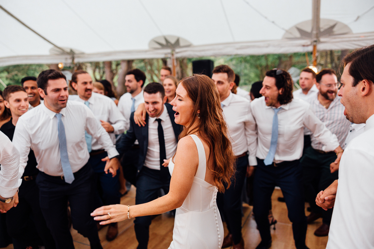 A woman in a white dress dances joyfully at a Seven Hills Inn wedding in Lenox MA, surrounded by guests in white shirts and ties under a tent. Everyone looks happy and engaged in the celebration.