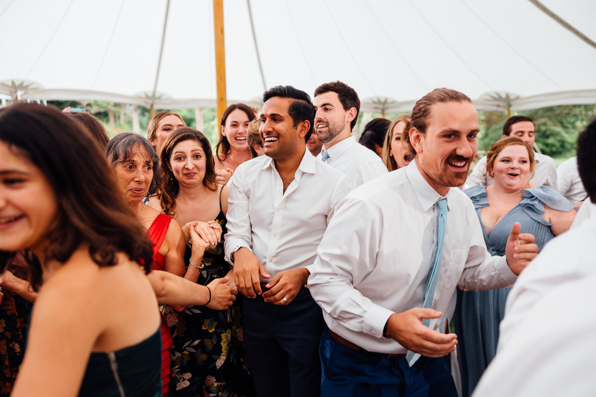 A group of people dressed in semi-formal attire laugh and dance together under a white tent, appearing joyful and energetic at a Lenox MA wedding celebration.