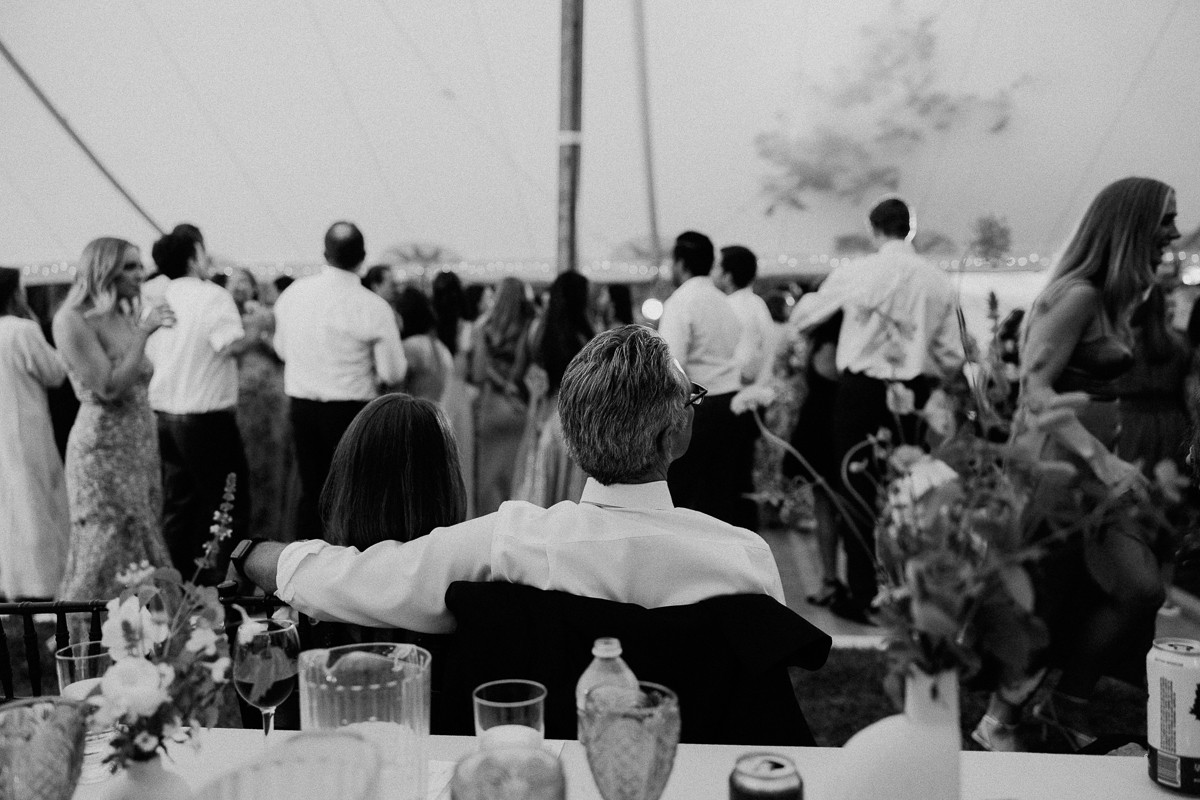 A black-and-white photo shows a couple seated at a table at a Seven Hills Inn wedding in Lenox, MA, facing away from the camera as they watch guests dance under a tent. Flowers and drinks are visible on the table in the foreground.