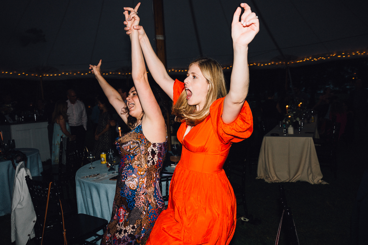 Two women joyfully dance with raised arms at a nighttime Seven Hills Inn wedding in Lenox MA, one in a bright orange dress and the other in a patterned dress, as tables and string lights glow under the tent.