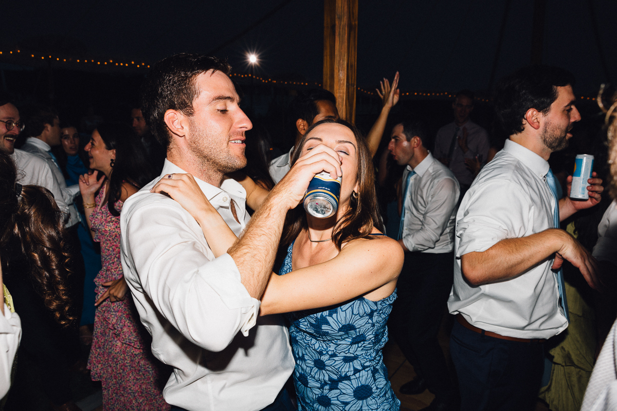 At a vibrant Seven Hills Inn wedding in Lenox, MA, a man and woman dance closely. The woman in a blue dress drinks from a can with a smile, surrounded by guests dancing and having fun in the background.