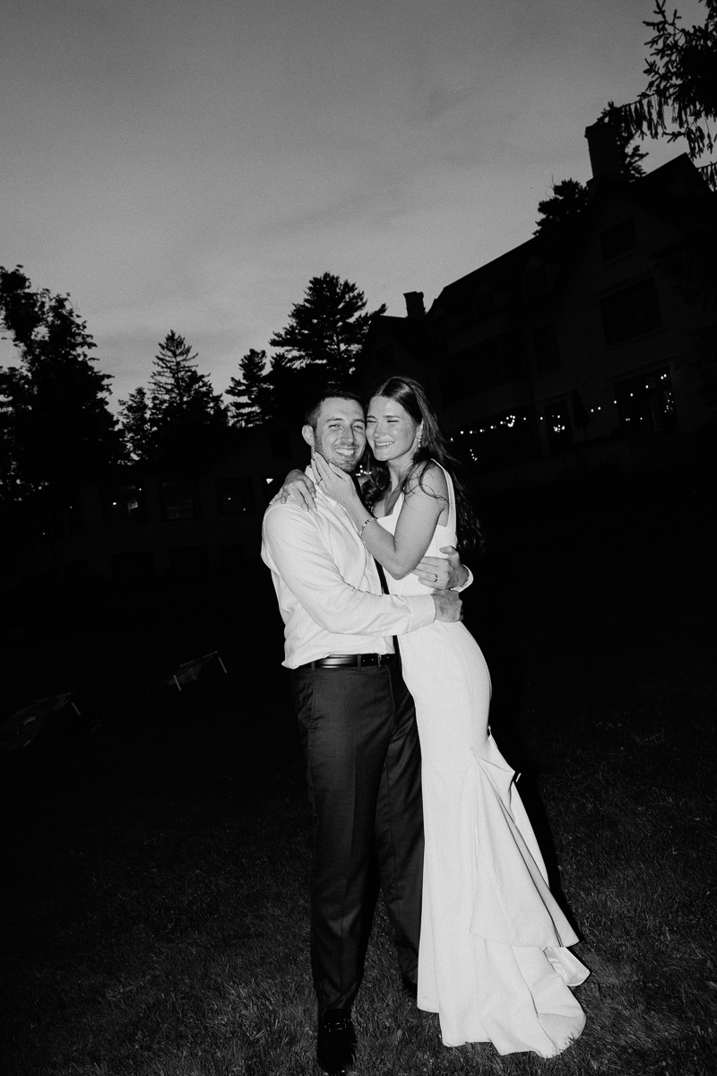 A couple dressed in formal attire, smiling and embracing outdoors at dusk, with trees and a large house silhouetted behind them. This black-and-white image captures a joyful moment from a Seven Hills Inn wedding in Lenox MA.