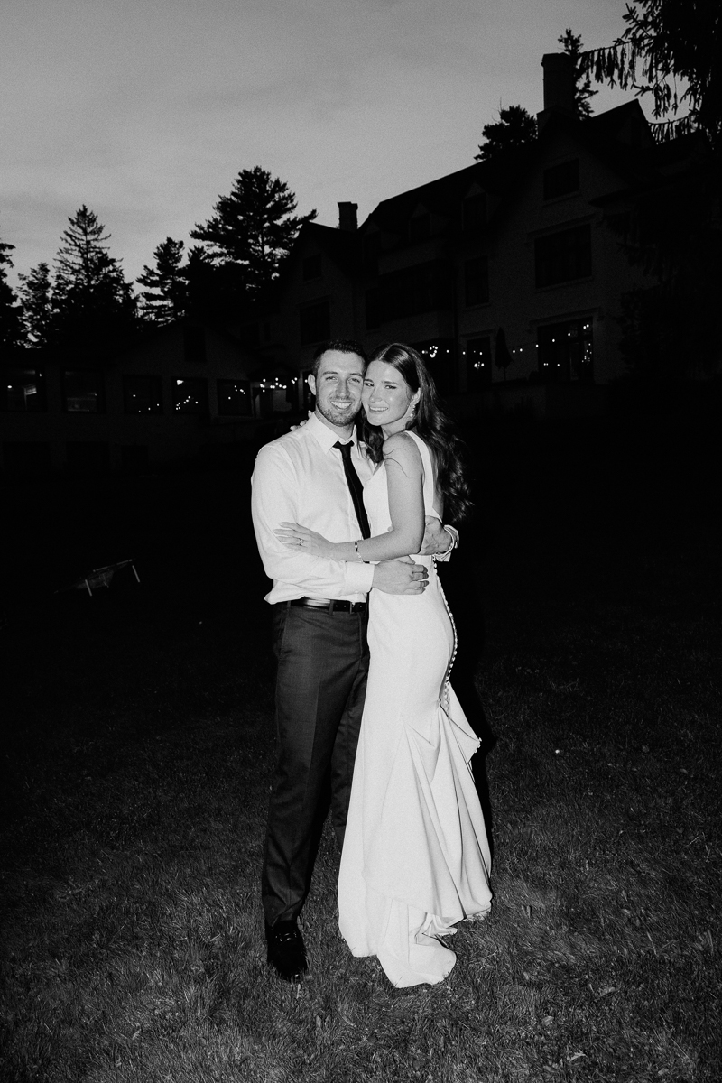 A couple dressed in formal attire, with the woman in a white gown and the man in a shirt and tie, embrace and smile outdoors at dusk in front of Seven Hills Inn during their Lenox MA wedding.
