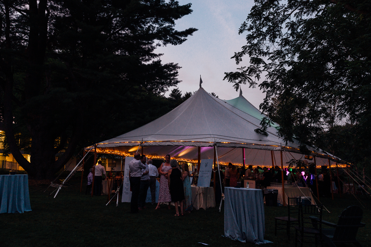 A large white event tent with string lights glows at dusk during a Seven Hills Inn wedding. People in formal attire mingle outside, surrounded by tall trees and tables with blue tablecloths on a grassy Lenox MA lawn.