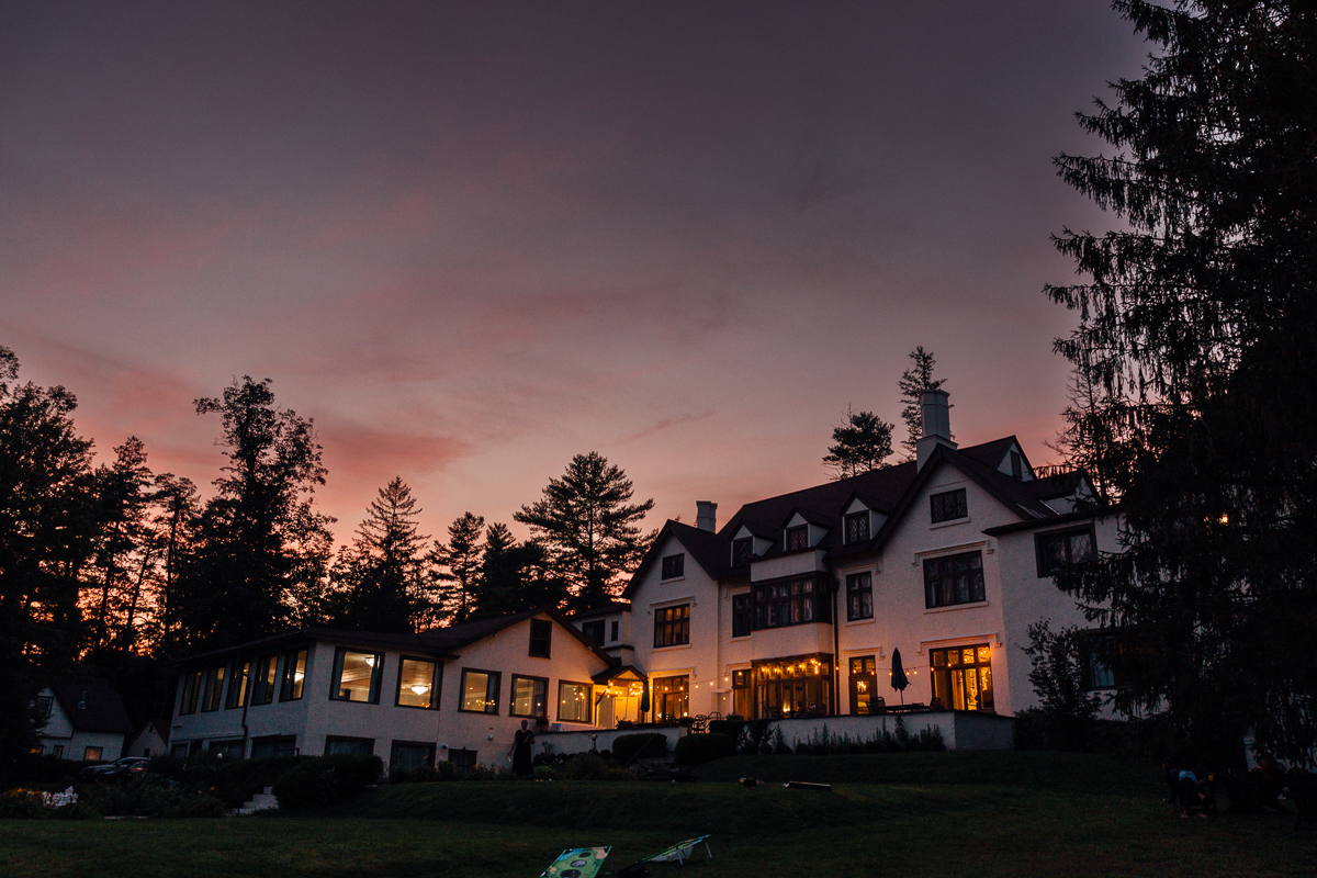 A large, white house with many windows is lit warmly from inside at dusk. Tall trees surround the Seven Hills Inn in Lenox, MA, and the sky glows purple and pink—a picturesque setting for a wedding evening.