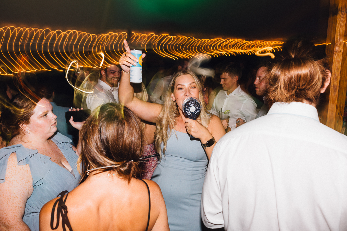 At a lively wedding at Seven Hills Inn in Lenox, MA, guests in party attire dance enthusiastically under string lights. One woman in front holds a drink and handheld fan, singing or cheering as the background lights create a dynamic blur.