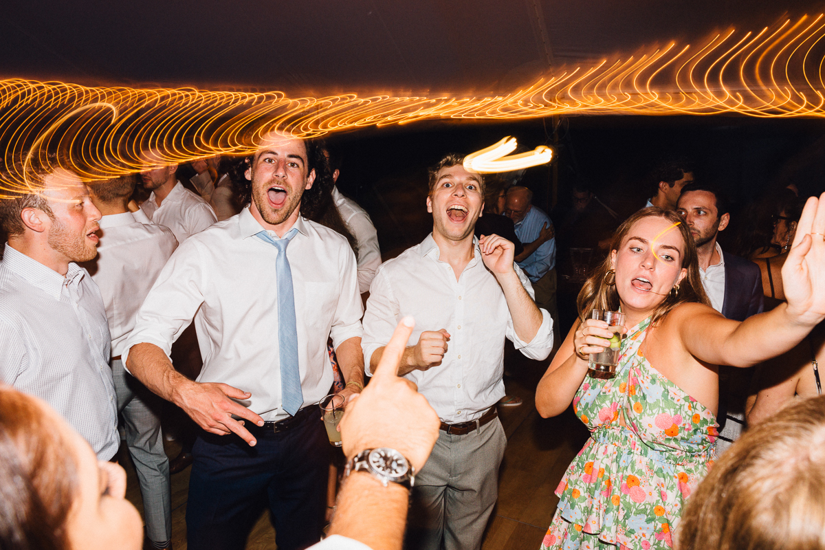 A group of people enthusiastically dancing and singing at a Seven Hills Inn Wedding in Lenox, MA, with a woman in a floral dress holding a drink and reaching out, as light trails create a dynamic effect above them.