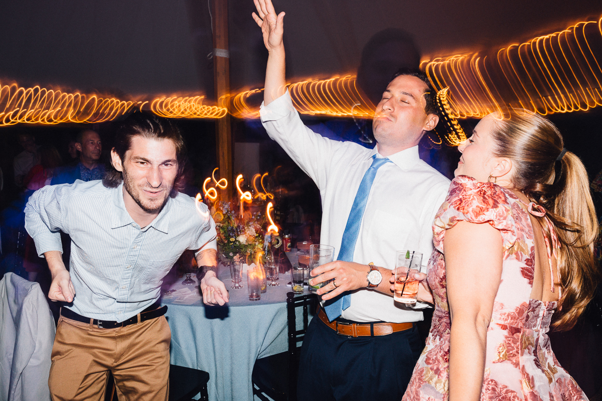 Three people enthusiastically dance at a Seven Hills Inn wedding in Lenox MA, holding drinks. Two men in dress shirts and a woman in a floral dress move energetically, with blurred lights creating a festive atmosphere in the background.