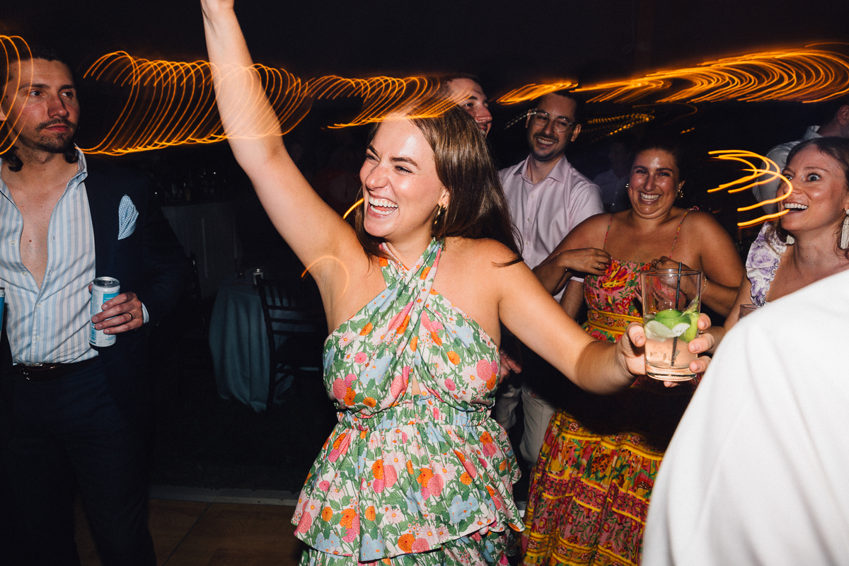 A woman in a floral dress smiles and raises her arm while holding a drink at a lively Seven Hills Inn wedding in Lenox MA. People around her laugh and dance, with blurred lights creating a festive atmosphere.