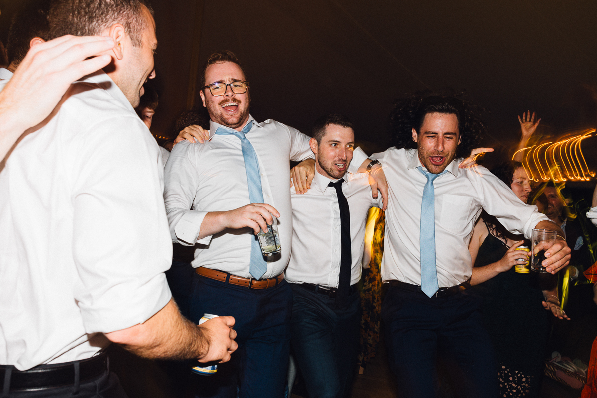 Four men in dress shirts and ties laugh and dance together at a lively Seven Hills Inn wedding in Lenox, MA, arms around each other, holding drinks as a festive crowd mingles under glowing string lights in the background.