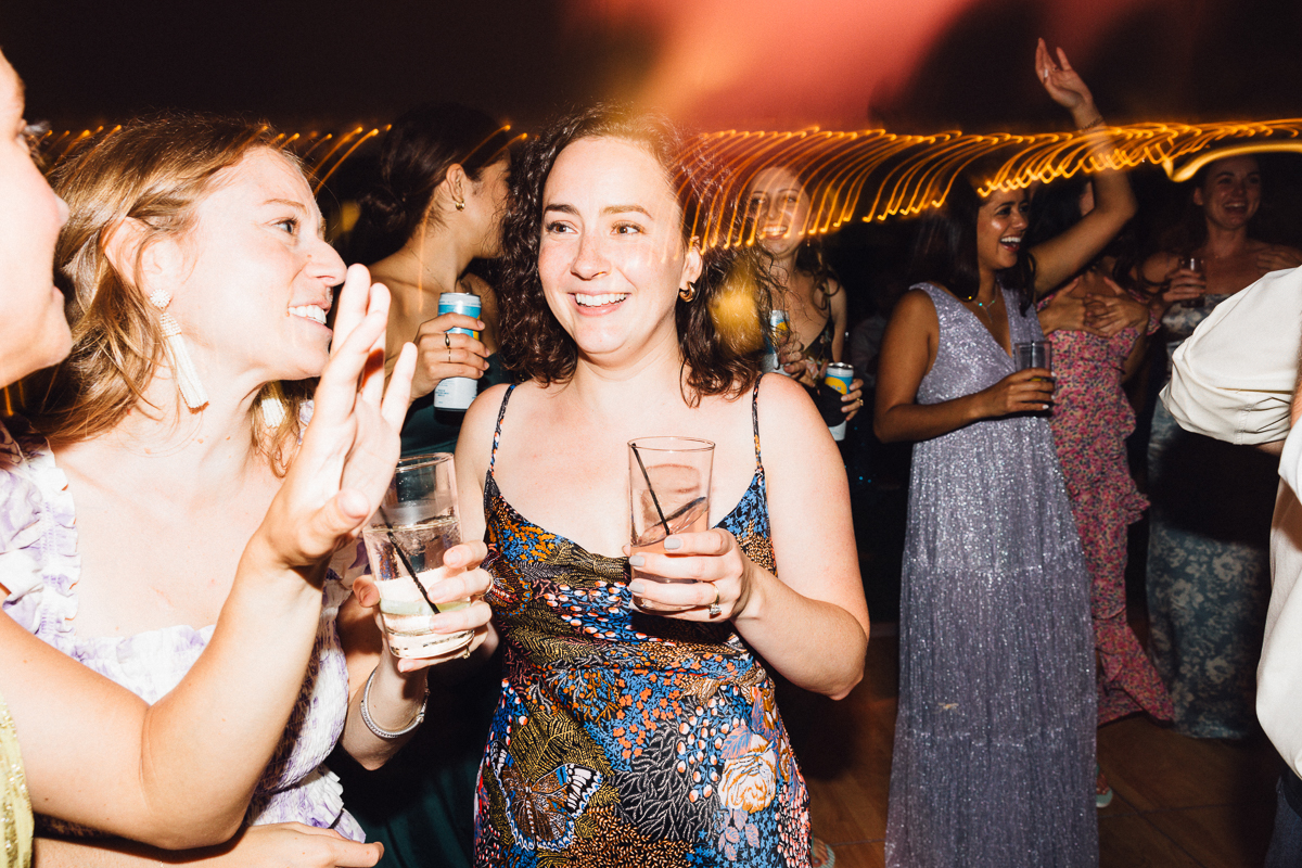 A group of women in colorful dresses laugh, smile, and hold drinks while dancing at a lively Seven Hills Inn wedding in Lenox MA, with blurred lights glowing in the background.