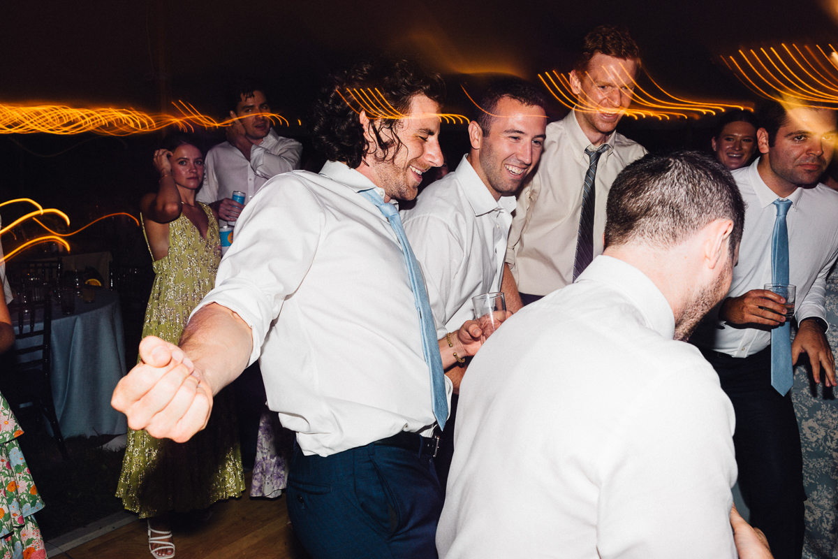 A group of people in dress shirts and ties are laughing and dancing energetically at a lively Seven Hills Inn wedding, with warm light trails in the background.