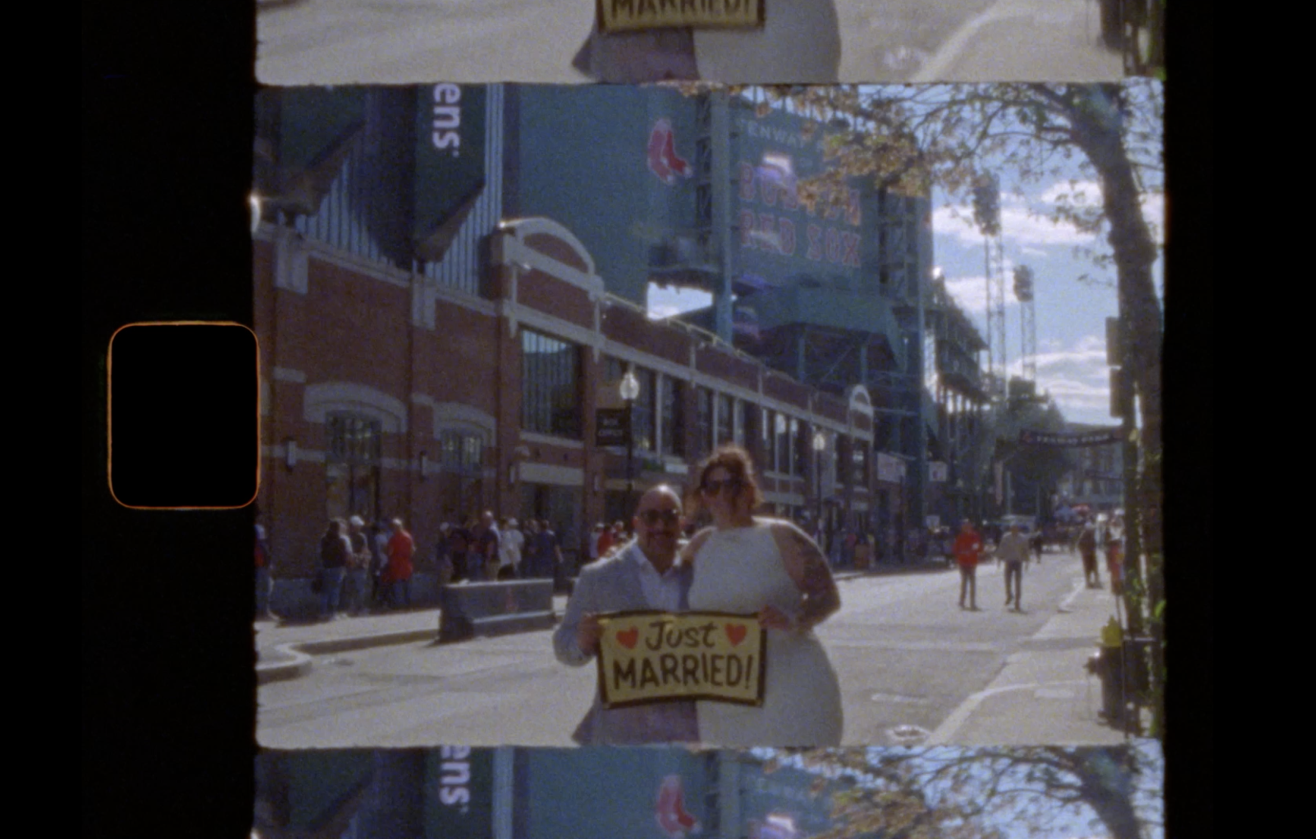 A couple stands on a city street holding a Just Married! sign. People and trees line the sidewalk, and a large stadium with Red Sox signage is visible in the background. The image has a vintage film look. It was taken on Super8 film by Boston Photographer Yaritza Colon