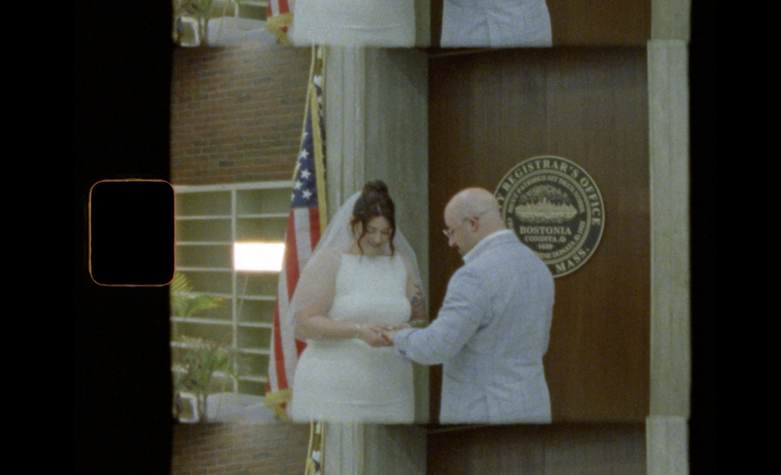 A bride and groom exchange rings in front of a Bostonia city seal and an American flag inside a courthouse. The bride wears a white dress and veil; the groom wears a light suit. The scene appears to be filmed on vintage film. It was on Super8 film by Boston Super8 photographer Yaritza Colon