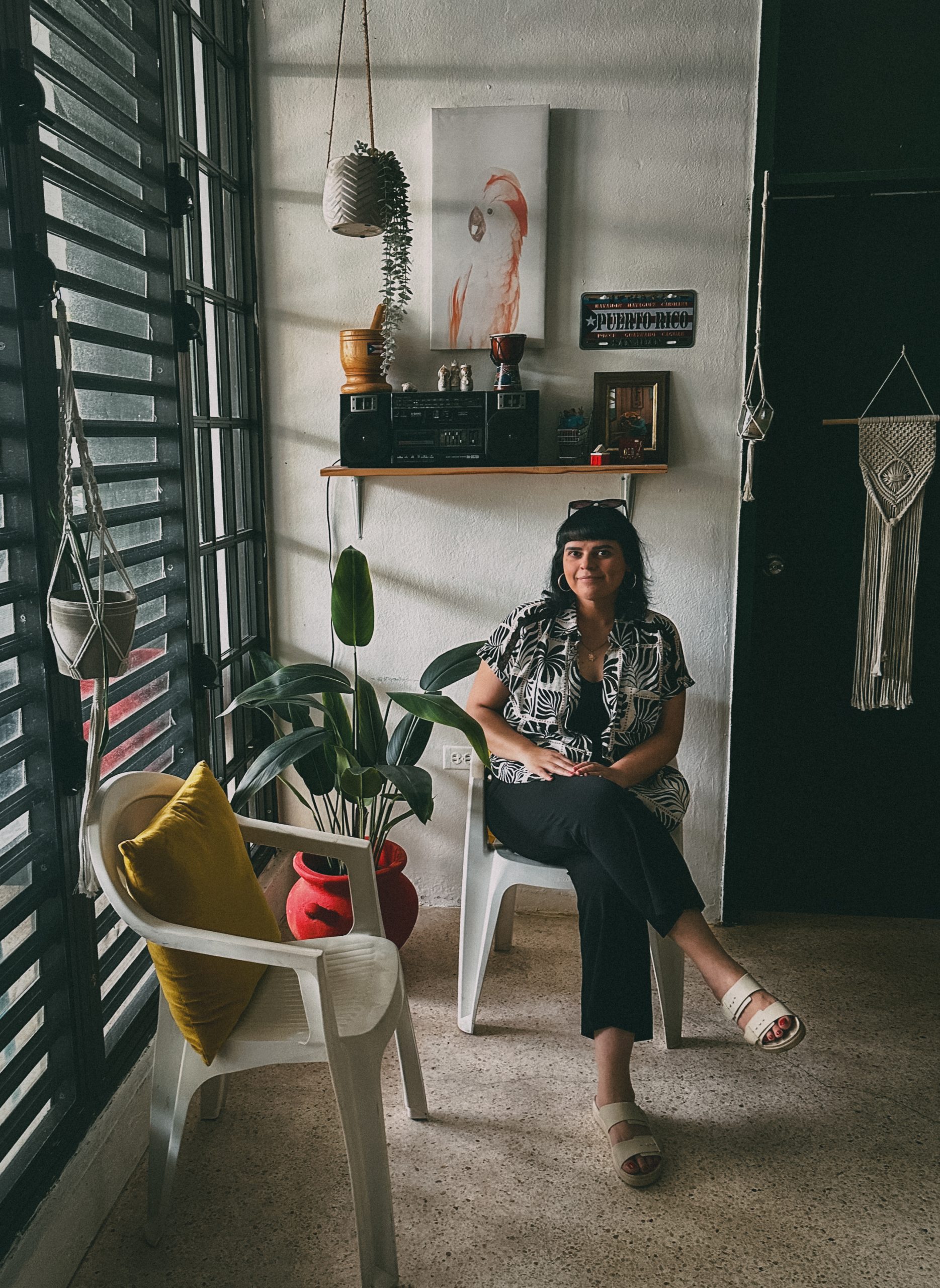 A woman sits on a white chair in a cozy, boho-style room with plants, wall art, and sunlight streaming through large windows. Another white chair with a yellow pillow is beside her.