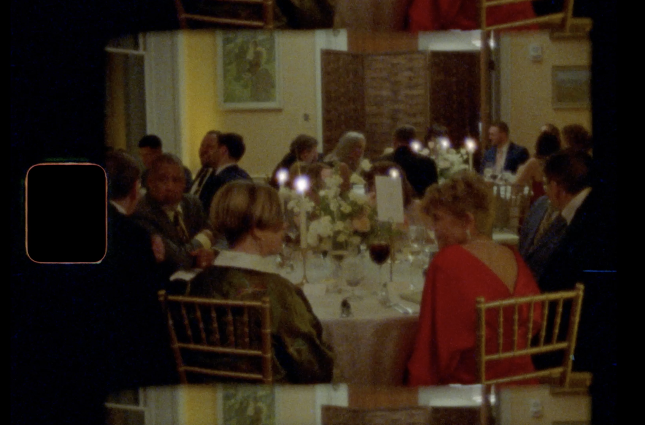 A dimly lit formal dinner with guests seated around round tables, flowers and candles as centerpieces, and gold chairs. The image has a grainy, vintage film effect with black film strip borders.